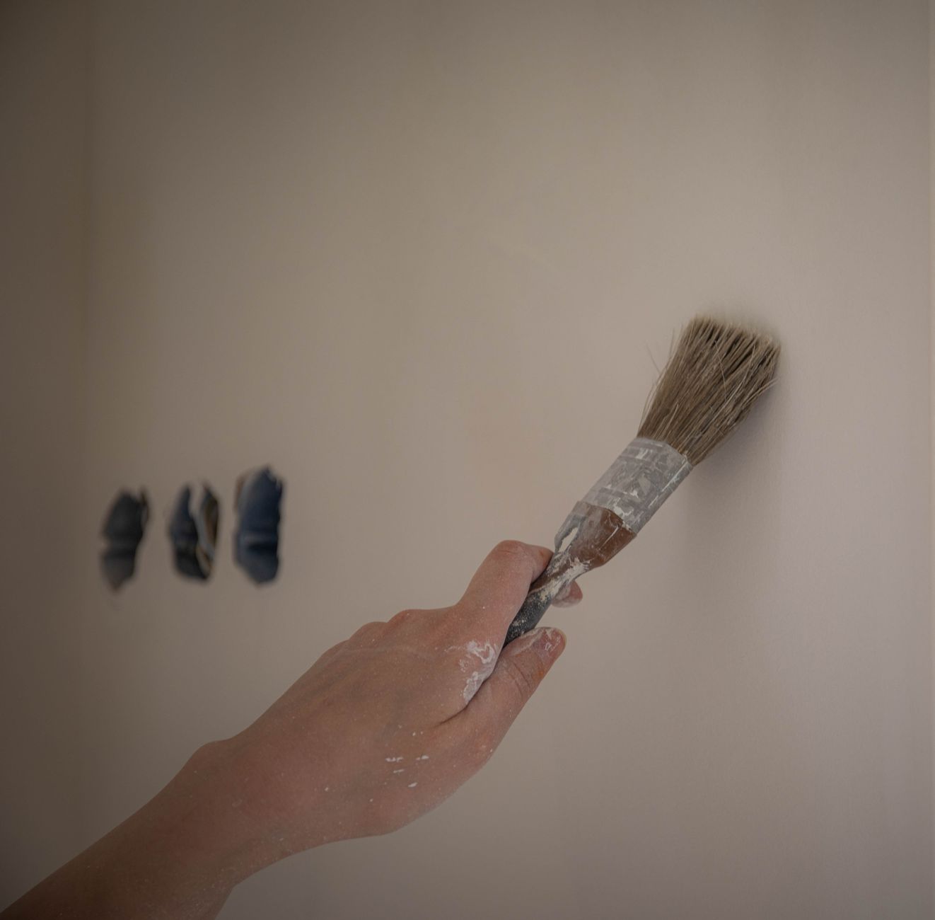 A close-up of a hand using a brush to paint a white wall, three color swatches visible in Temecula, CA
